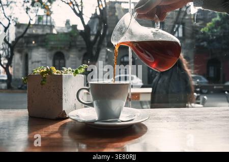 Hand of Hispanic woman pouring black coffee into white porcelain cup on wooden table in front of glass wall in cafe Stock Photo