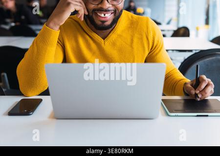 Black man with diastema wearing yellow sweater working on digital pen and tablet while sitting at desk with laptop and smartphone in office Stock Photo