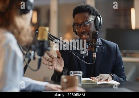 Black man and caucasian woman wearing headphones having conversation while sitting at round table with microphones recording podcast in studio Stock Photo