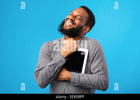 Black man with closed eyes hugging tablet while standing against blue background in studio Stock Photo
