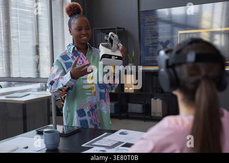 Black female programmer with high bun standing and presenting new VR glasses to her colleagues during team meeting in office Stock Photo