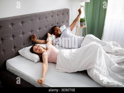 Black and caucasian couple waking up stretching their arms while lying on bed at home Stock Photo