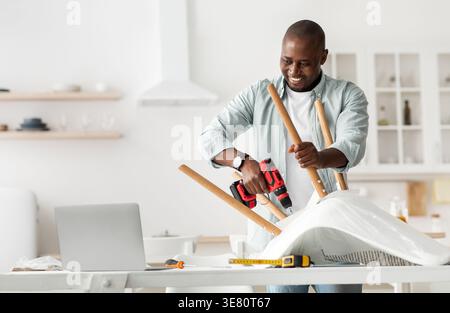 Black man using drill assembling chair watching on laptop in kitchen at home Stock Photo