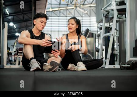 Asian man wearing sportswear using tablet talking to female friend while sitting on floor together in gym Stock Photo