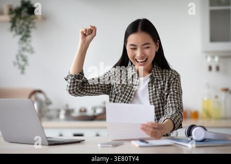 Cheerful Asian woman with clenched fist reading letter while sitting at table with headphones and laptop at home Stock Photo