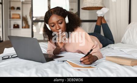 Black woman with curly hair wearing headphones taking notes and using laptop while lying down on bed in bedroom at home Stock Photo