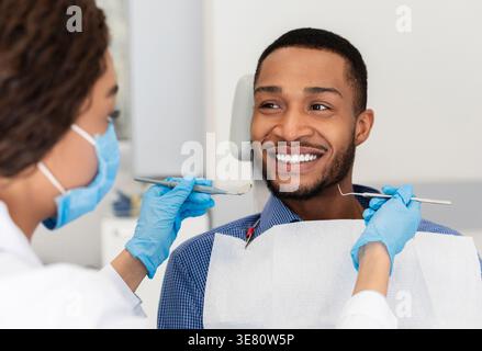 Black male patient getting his teeth checked by female dentist wearing gloves and face mask while sitting on chair in clinic Stock Photo