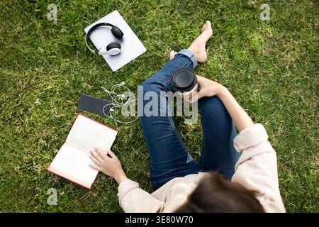 Hispanic female college student holding disposable coffee cup sitting with crossed legs on grass near book, laptop and headphones in campus park du... Stock Photo