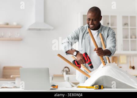 Black man using drill assembling chair on table standing in kitchen at home Stock Photo