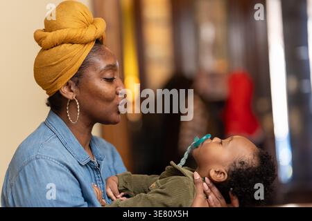 Black mother wearing yellow head wrap and long sleeve denim shirt holding and playing with her baby boy with pacifier while sitting at home Stock Photo
