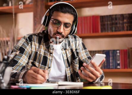 Indian male student with headphones wearing eyeglasses holding smartphone while writing down notes sitting at table in library Stock Photo