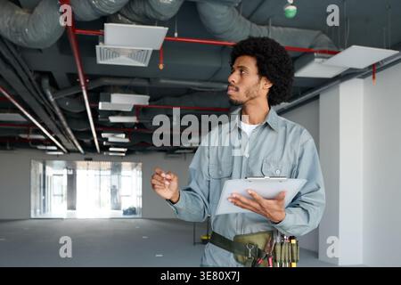 Black male inspector in gray uniform holding clipboard and looking away while standing and checking building under construction Stock Photo