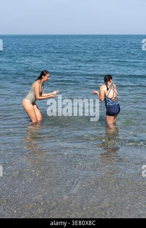 Hispanic female friends in bikinis splashing water at each other while standing at beach on sunny day Stock Photo