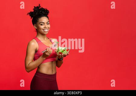 Black woman holding bowl of salad and fork with green bell pepper standing against red background in studio Stock Photo
