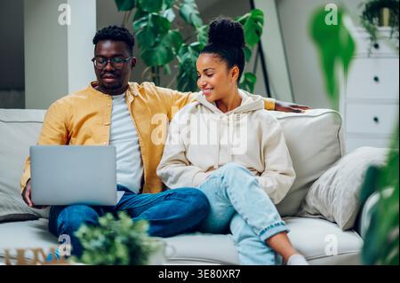 Black man using laptop while sitting on sofa beside mixed-race woman having coffee at home Stock Photo