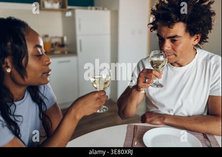 Black and mixed-race couple sitting at table drinking wine having romantic dinner together in kitchen at home Stock Photo