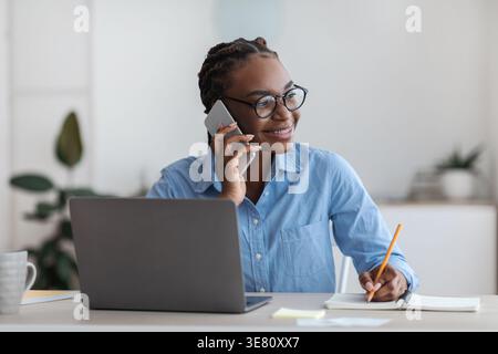Black business woman with braids wearing eyeglasses having phone call taking notes while sitting at table with laptop in home office Stock Photo