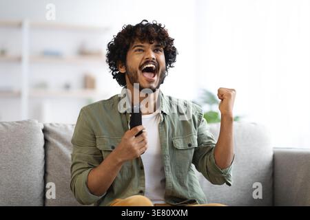 Cheerful Asian man with curly hair holding tv remote and showing clenched fist while sitting on sofa in living room at home Stock Photo