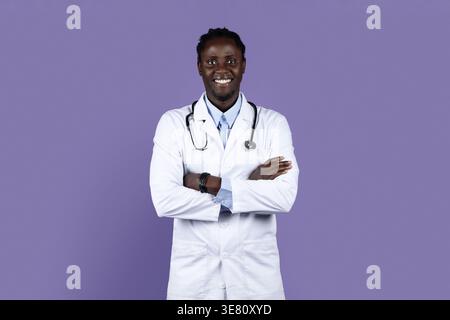 Smiling Black male doctor with dreads wearing white suit with stethoscope standing with arms crossed against purple background in studio Stock Photo
