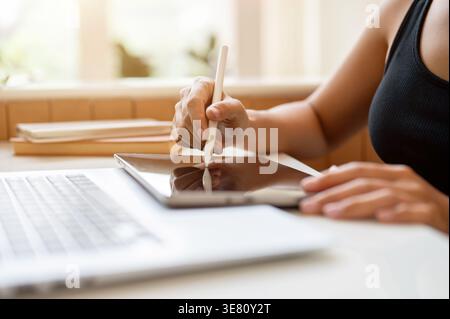 Hands of Thai woman writing on tablet using stylus pen while sitting at table with laptop in cafe Stock Photo