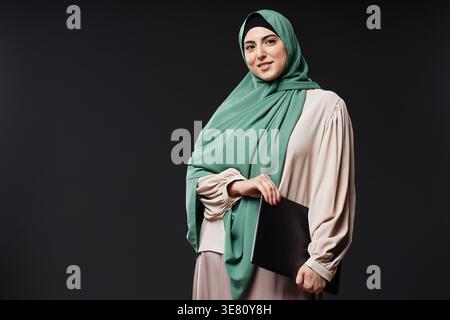 Smiling mixed-race woman wearing hijab carrying laptop while standing against black wall in studio Stock Photo