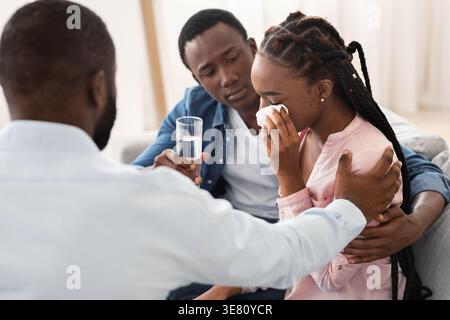 Black man giving water and comforting his wife having mental health problems while sitting on couch and talking with male therapist in office Stock Photo