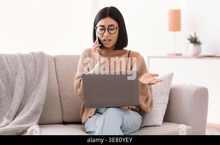 Asian woman wearing eyeglasses having phone call while working on laptop sitting on sofa in living room at home Stock Photo