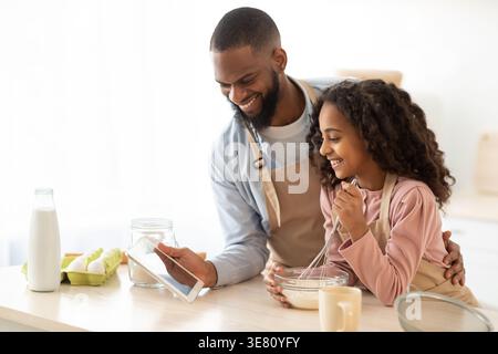 Black man wearing apron sharing tablet with daughter mixing ingredients for baking in bowl using whisk while standing close together at table in ki... Stock Photo