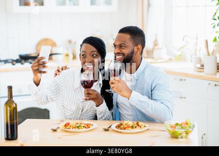 Black couple holding glasses of wine taking selfie using smartphone sitting at table in kitchen at home Stock Photo