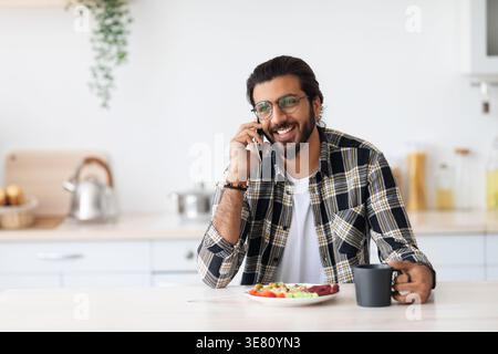 Arab man wearing eyeglasses having phone call and eating breakfast looking away while sitting at table in kitchen at home Stock Photo