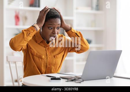 Shocked Black woman with short hair looking at laptop screen and touching her head while sitting at table and looking at laptop screen in office Stock Photo