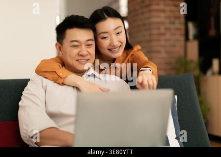 Asian woman embracing husband from behind sitting on sofa using laptop together in living room at home Stock Photo