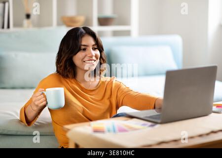 Smiling Hispanic woman holding coffee cup while working on laptop sitting on floor in front of bed in bedroom at home Stock Photo