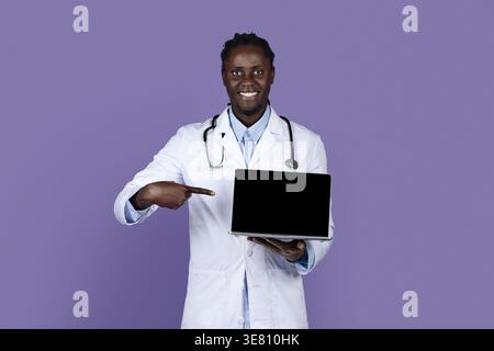 Smiling Black male doctor with dreads wearing white suit with stethoscope pointing at laptop on hand while standing against purple background in st... Stock Photo