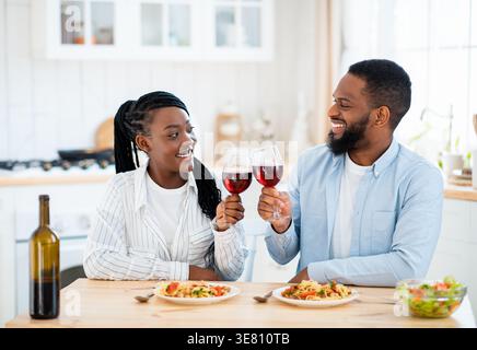 Black couple looking at each other toasting glasses of wine having date sitting at table in kitchen at home Stock Photo