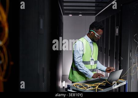 Black male network technician wearing protective glasses and vest standing and repairing servers in data center Stock Photo