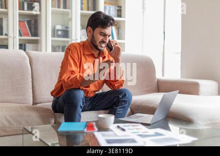 Disappointed Middle Eastern man gesturing hand looking at laptop while having phone call sitting on couch at home Stock Photo