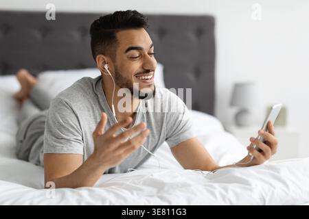 Arab man in earphones having video call using smartphone while lying on bed in bedroom at home Stock Photo