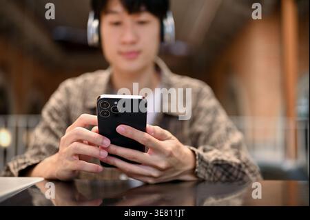Asian man wearing headphones using smartphone while sitting at table in coffee shop Stock Photo