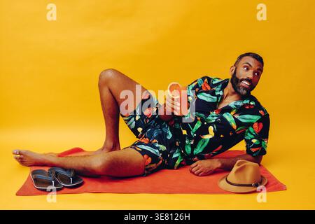 Mixed-race bearded man leaning on elbow holding glass of orange juice and looking away while lying sideways on towel with flip flops and fedora hat... Stock Photo