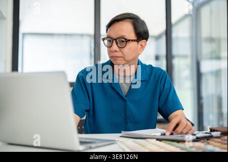Asian middle-aged male interior designer looking at laptop sitting at the table with materials in office Stock Photo