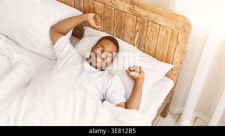 Smiling Black man in white shirt stretching arms with closed eyes while lying on bed at home Stock Photo