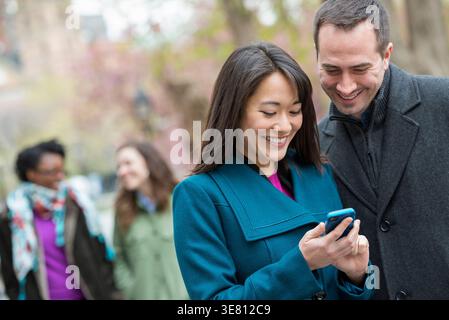 Korean woman showing smartphone to caucasian male friend standing in park during daytime Stock Photo