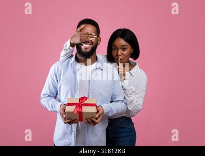 Black woman covering her boyfriend's eyes and making hush gesture while standing against pink background in studio Stock Photo