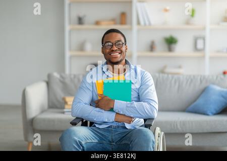 Smiling Black disabled man wearing eyeglasses embracing notebook while sitting on wheelchair in living room at home Stock Photo