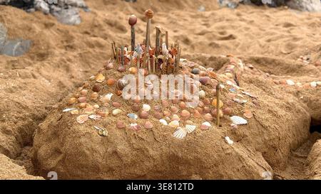 Intricate sand sculpture decorated with numerous sea shells and wooden sticks on a beach shoreline Stock Photo