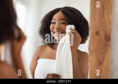 Smiling Black woman with curly hair wrapped in bath towel drying face after shower looking at mirror while standing in bedroom at home Stock Photo