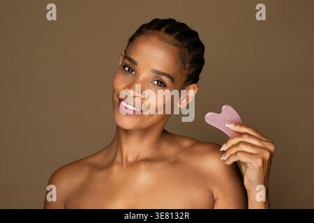Black woman holding Gua Sha quartz stone scraper while standing against brown background in studio Stock Photo