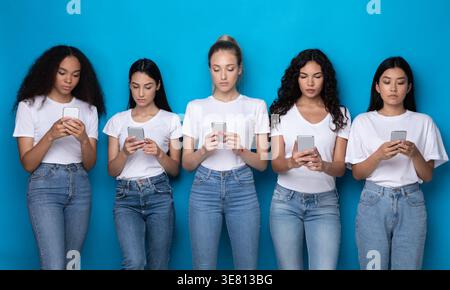 Diverse group of women wearing white shirts and jeans using smartphones standing against blue background in studio Stock Photo