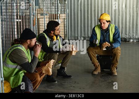 Mixed-race male worker wearing safety hat and vest talking to male colleagues having lunch during break sitting in warehouse Stock Photo